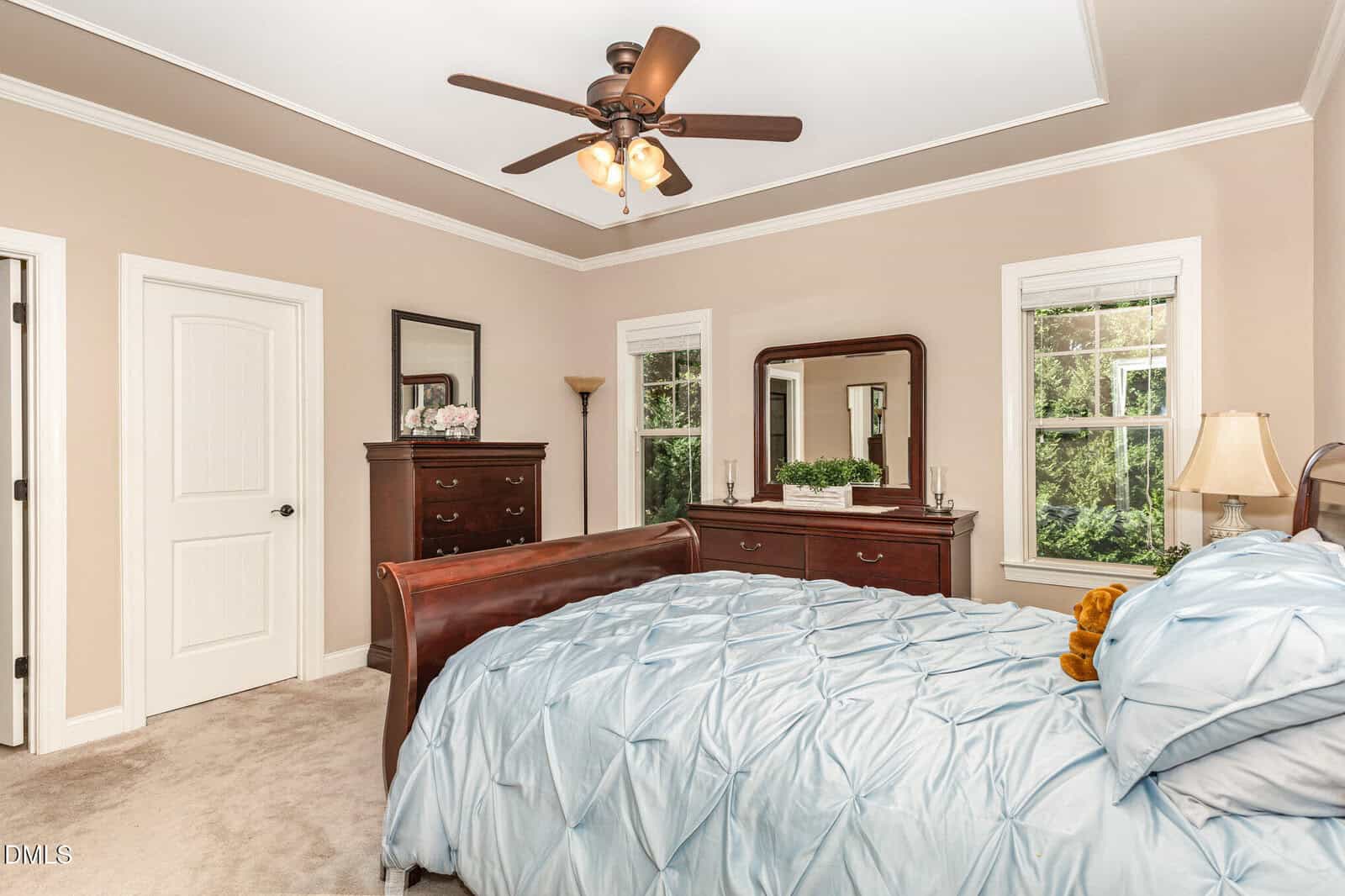 A bedroom with beige walls, a ceiling fan, light carpet, a bed with a blue comforter, two dressers with mirrors, and two windows showing greenery outside. A lamp and decorative items are on the dressers.