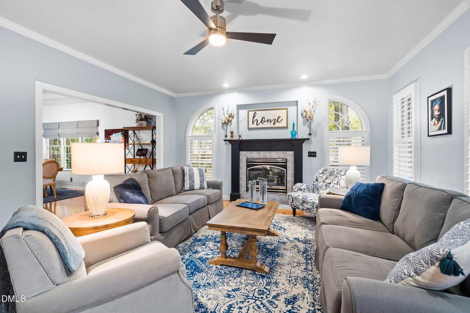 Bright living room with two gray sofas, an armchair, a wooden coffee table, blue patterned rug, fireplace with "home" sign above, arched windows, and light blue walls with white trim.