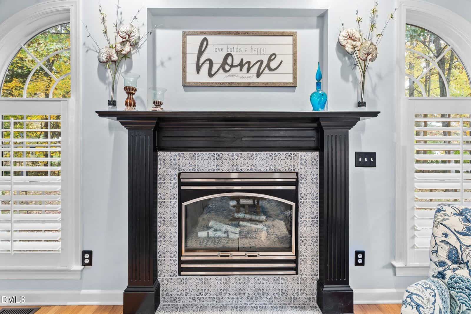 Black fireplace with patterned tile surround, flanked by windows with white shutters. Decor includes vases, a blue glass bottle, and a framed sign that reads, “Love builds a happy home.” Light blue walls and wood floor.