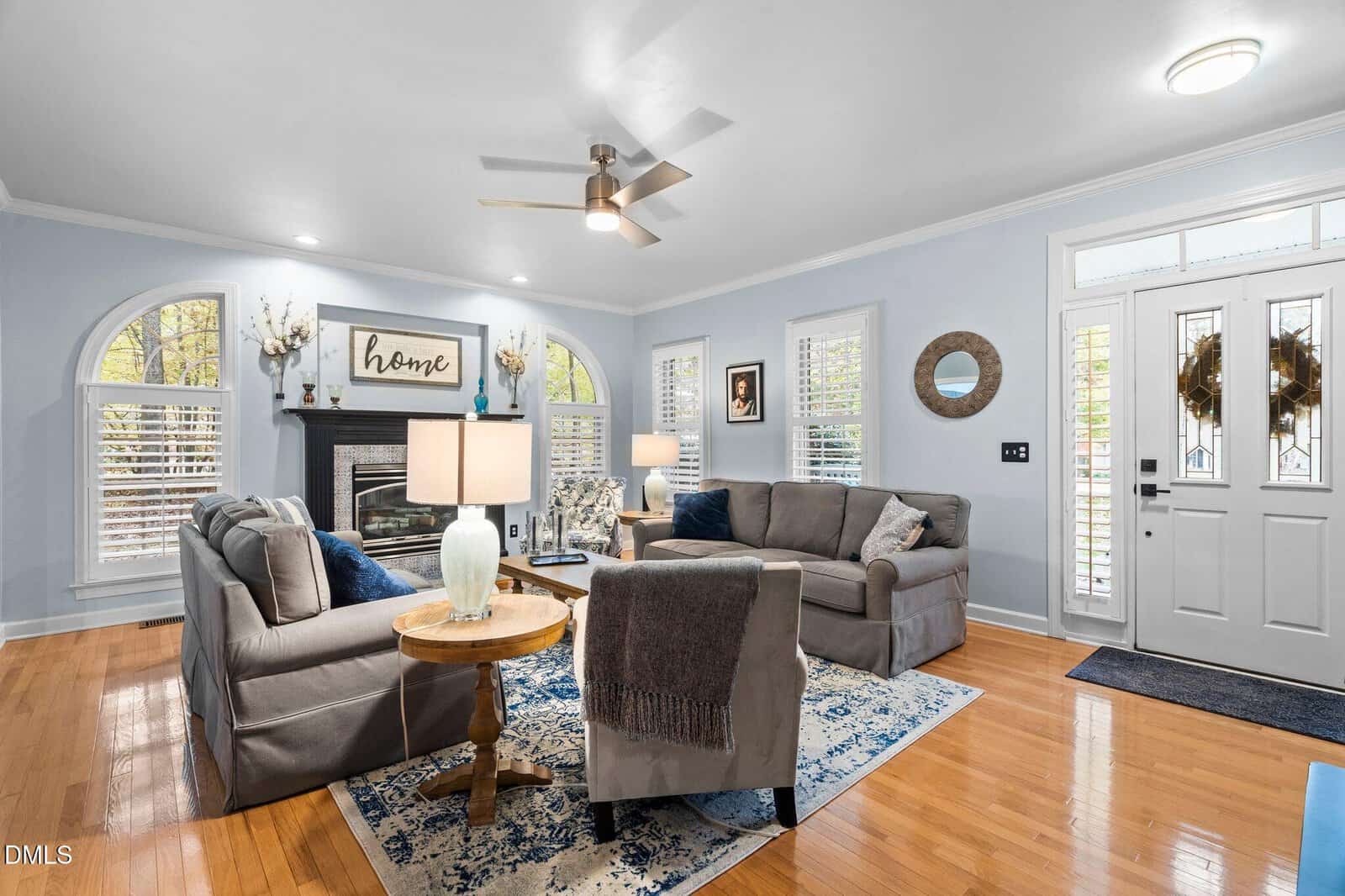 Bright living room with two gray sofas, an armchair, a fireplace, lamps, and a decorative “home” sign. Hardwood floors, blue accents, large windows, and a glass-paneled front door create a cozy, inviting space.