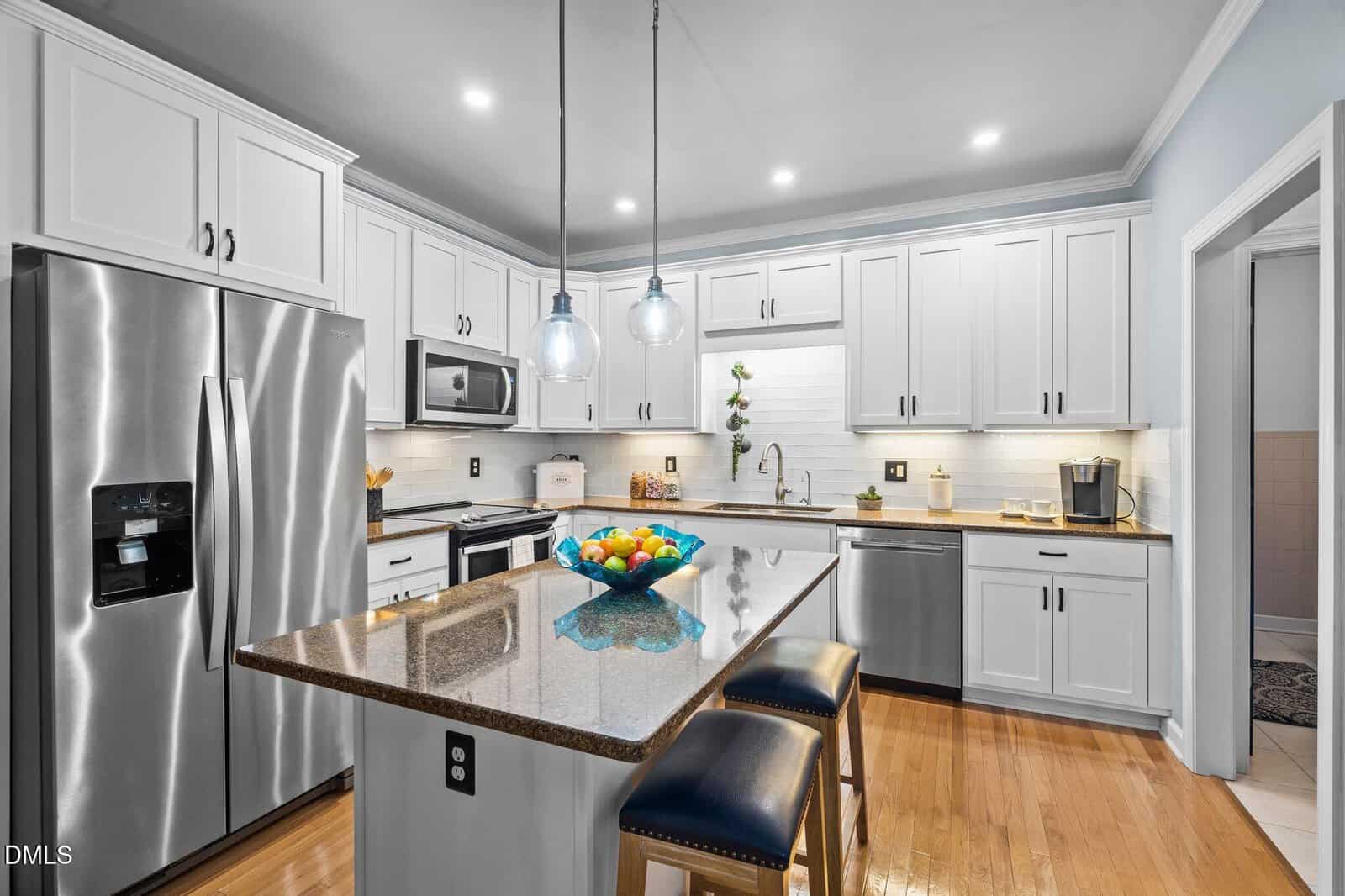 Modern kitchen with white cabinets, stainless steel appliances, a center island with two stools, pendant lights, and a bowl of fruit. Light wood floor and neutral backsplash complete the bright, clean look.
