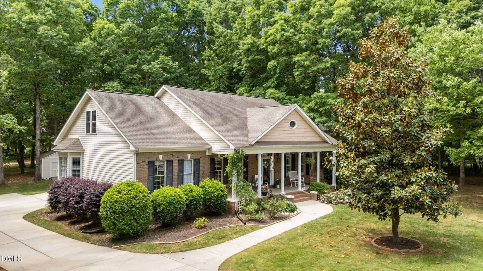 A beige, single-story house at 120 Georgetown Woods Drive, Youngsville, features a front porch with two rocking chairs, surrounded by green bushes and trees. A curved driveway leads to the home, and a large tree stands in the front yard.