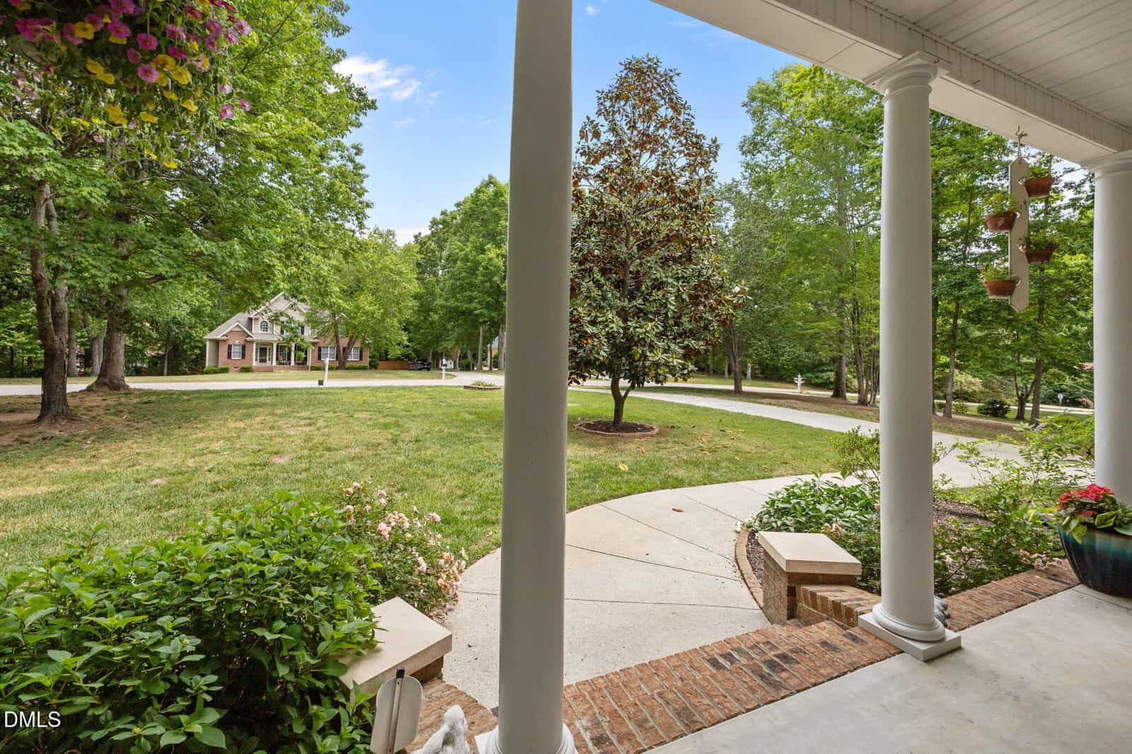View from a white-columned porch overlooking a curved walkway, green lawn, trees, and a two-story house with a gabled roof across the driveway on a sunny day.