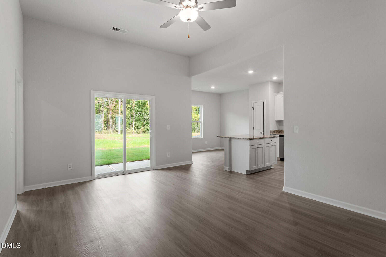 A spacious, unfurnished living area with light wood flooring, white walls, a ceiling fan, sliding glass doors leading to a patio, and an open view into a modern kitchen with white cabinets and granite countertops.