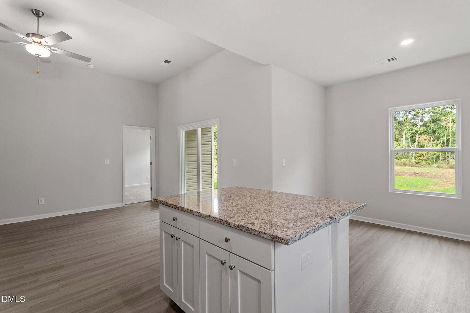 A modern, empty room with light gray walls, wood flooring, a ceiling fan, and windows letting in natural light. A kitchen island with a granite countertop and storage cabinets is in the foreground.