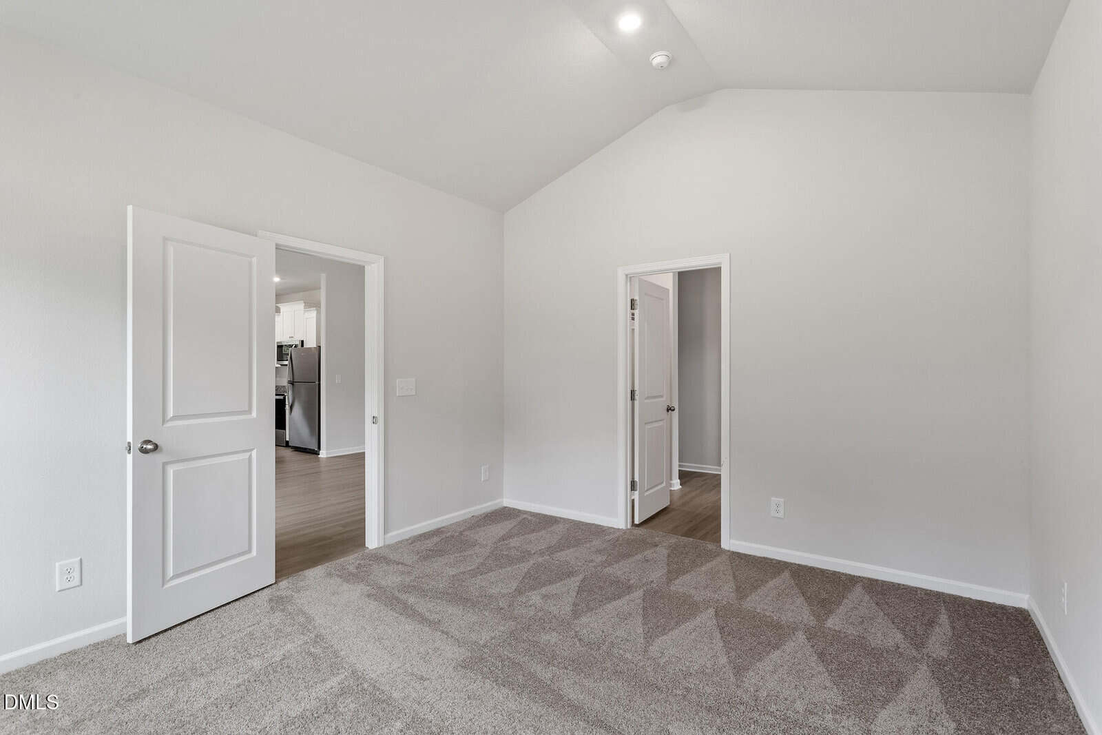 A carpeted, empty room with light gray walls, double doors open to a hallway, and a single door leading to another room. The ceiling is vaulted and a portion of a kitchen is visible through the open doors.