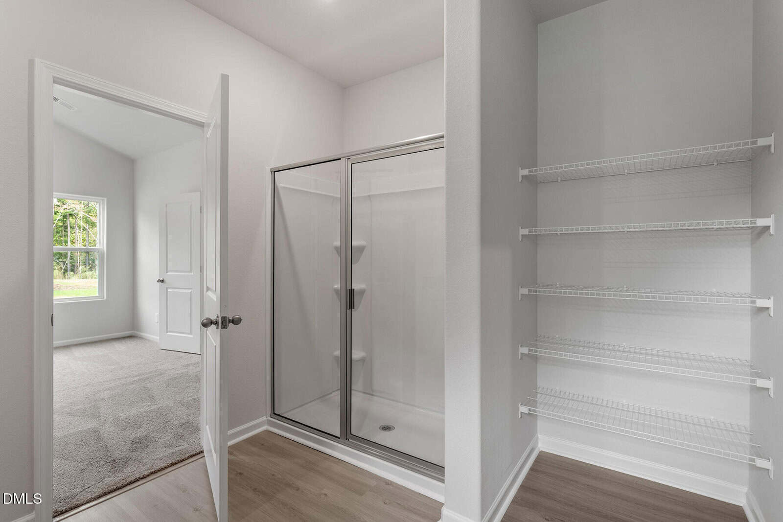 A bathroom with a glass shower, white walls, wood floor, and an open door leading to a carpeted bedroom. There are white wire shelves on the right side of the image.