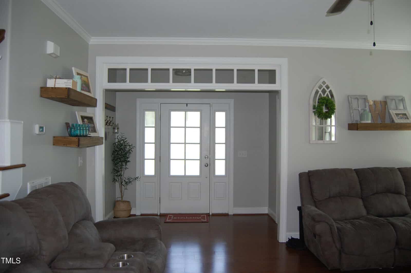 A living room with gray walls, a brown sectional sofa, floating wooden shelves with decor, a potted plant, and a white front door with glass panels and sidelights. Hardwood floors and crown molding are visible.