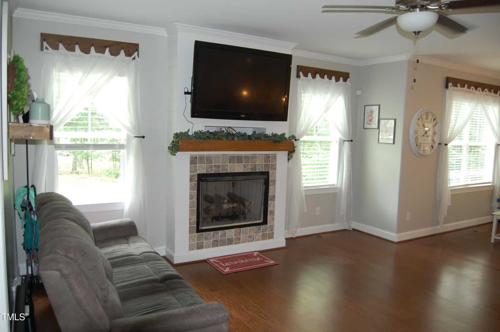 A living room with hardwood floors, a gray couch, a fireplace with a TV mounted above it, and several windows with white curtains letting in natural light. There is a ceiling fan and wall decorations.
