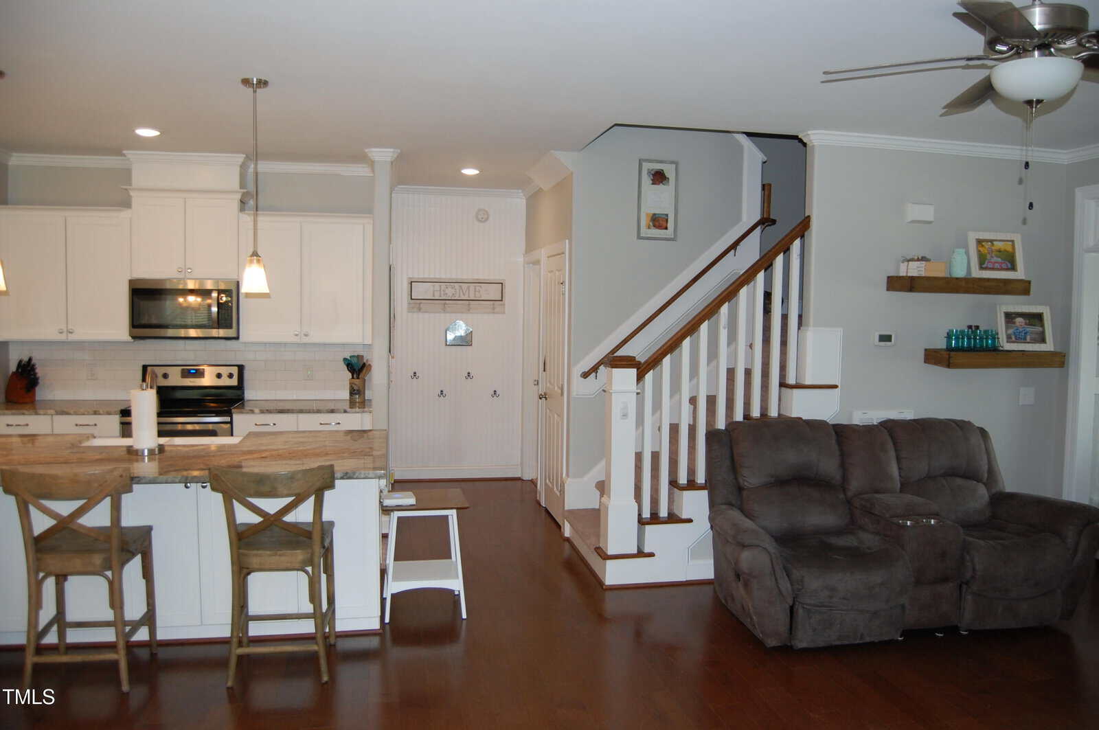 Open-concept kitchen and living area with white cabinets, stainless steel appliances, two barstools at a kitchen island, brown recliner seating, and a staircase with wooden handrails leading to the upper floor.