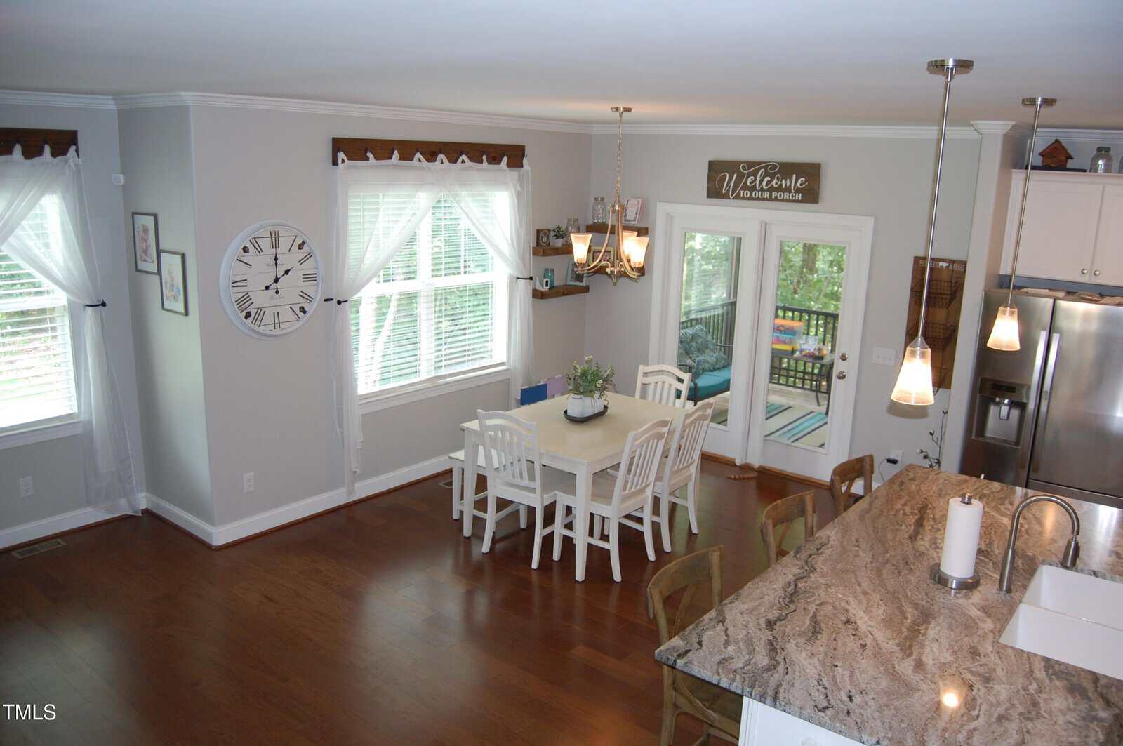 A bright kitchen and dining area with wood floors, white cabinetry, a marble countertop, a rectangular dining table with four chairs, large windows with white curtains, and a chandelier above the table.
