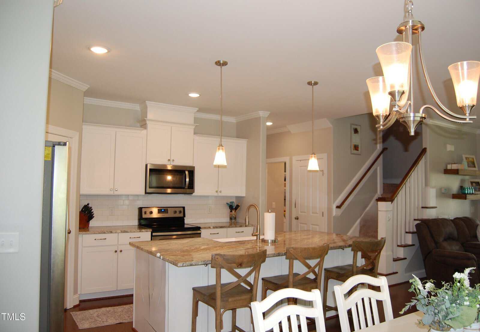 Modern kitchen with white cabinets, stainless steel appliances, an island with a granite countertop, three bar stools, pendant lights, and a view of the living area and staircase in the background.