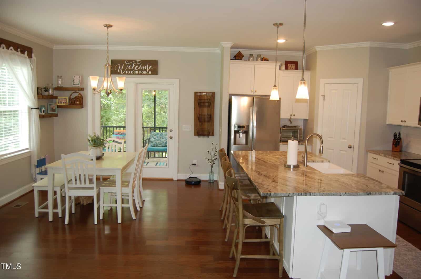 A modern kitchen with white cabinets, stainless steel appliances, a large island with barstools, pendant lighting, and a dining area with a table and chairs near windows and a glass door leading outside.