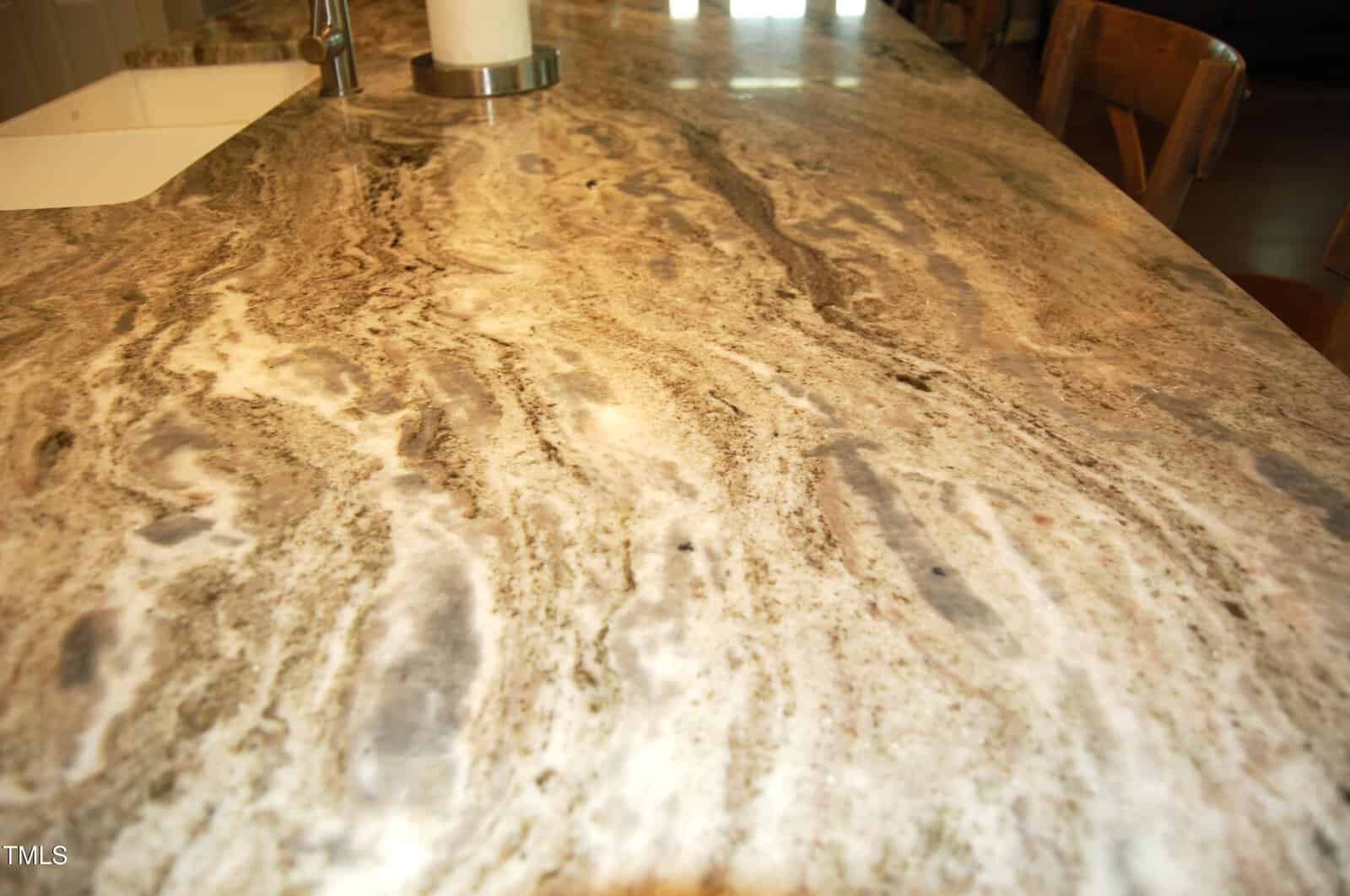 A close-up view of a kitchen countertop with a beige, brown, and gray marbled pattern. A white sink and a paper towel holder are partially visible in the background.