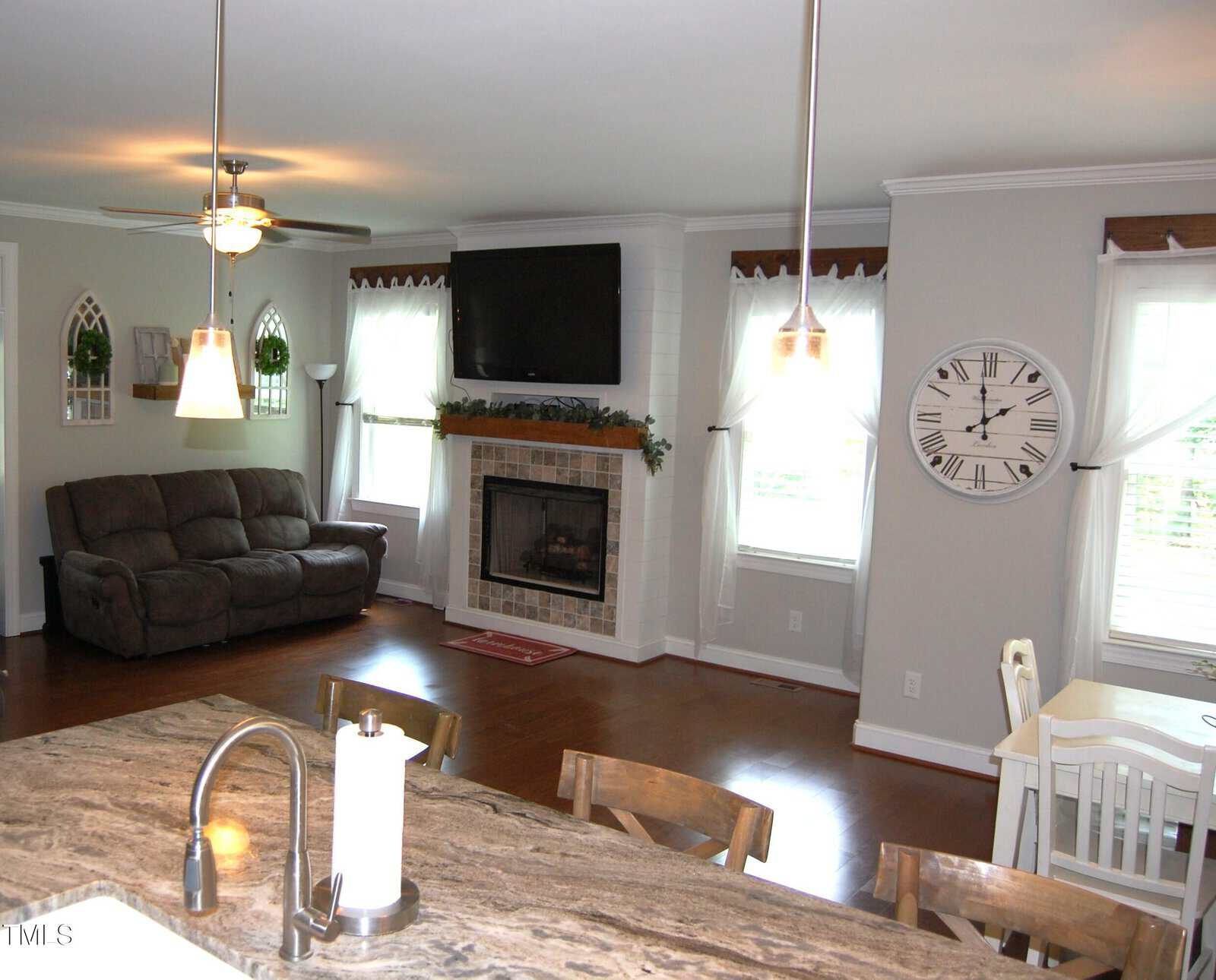 A living room with hardwood floors, a brown sofa, a wall-mounted TV above a fireplace, large windows with sheer curtains, a wall clock, and a kitchen island with a sink and paper towel holder in the foreground.