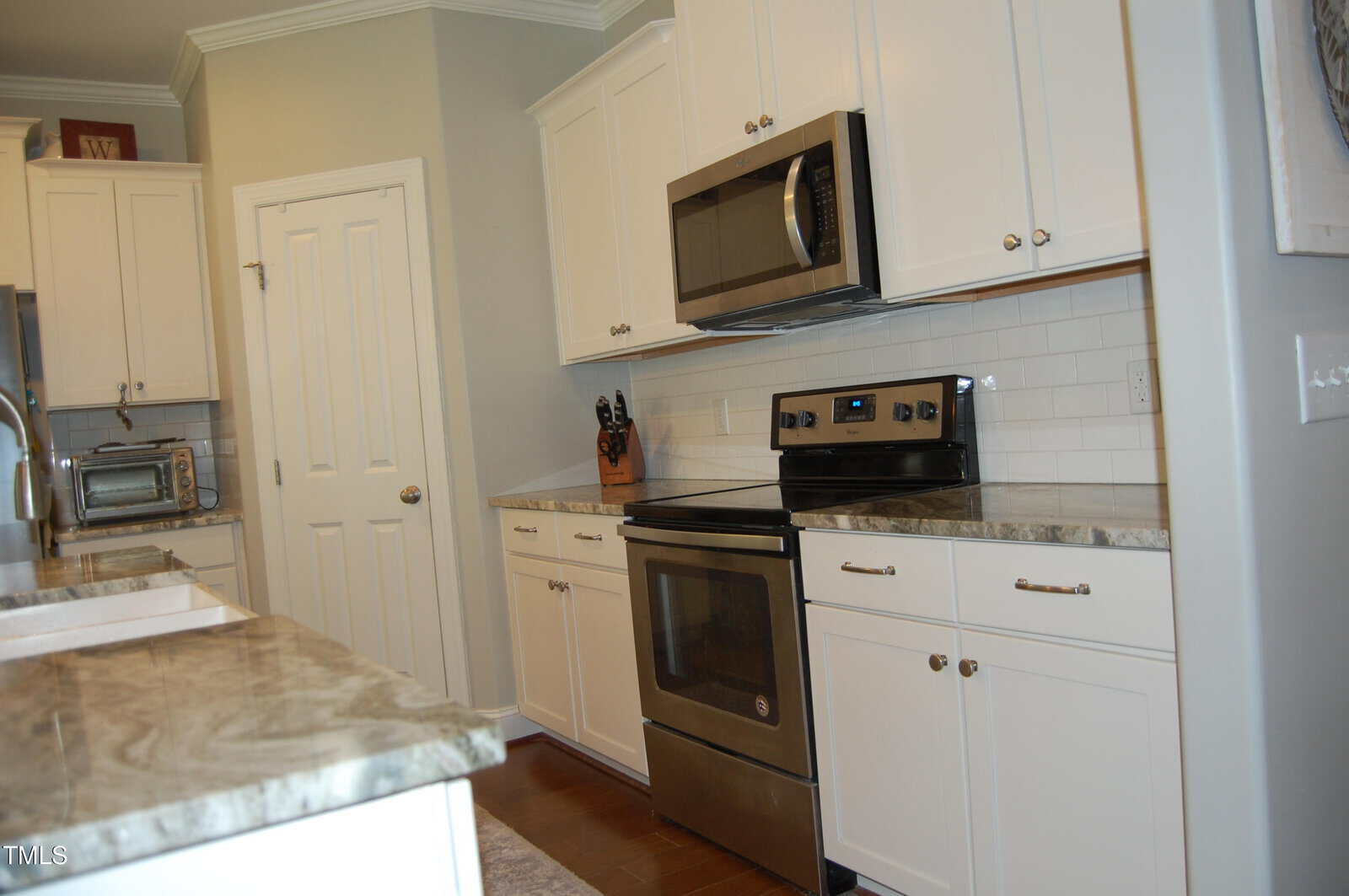 Modern kitchen with white cabinets, stainless steel oven, microwave, and marble countertops. Knife block and toaster are on the counters. Subway tile backsplash and wood floor are visible.