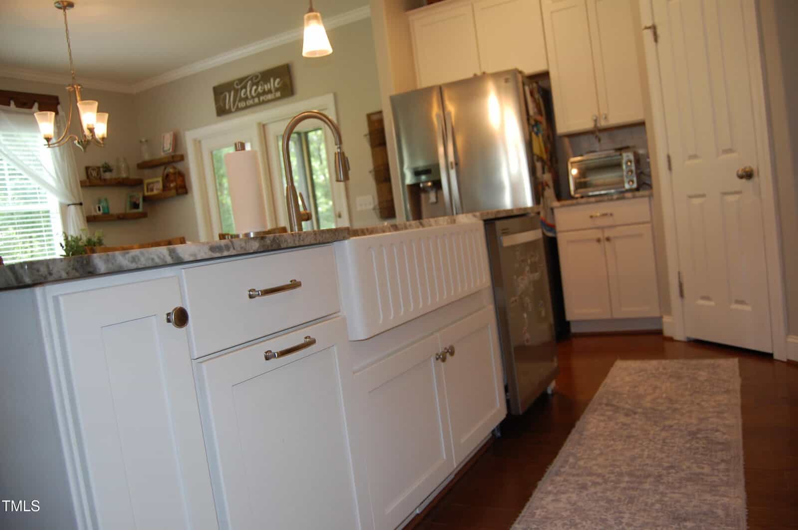 A modern kitchen with white cabinets, a farmhouse sink, stainless steel appliances, wooden floors, and a gray rug. Sunlight streams in through a window with sheer curtains.