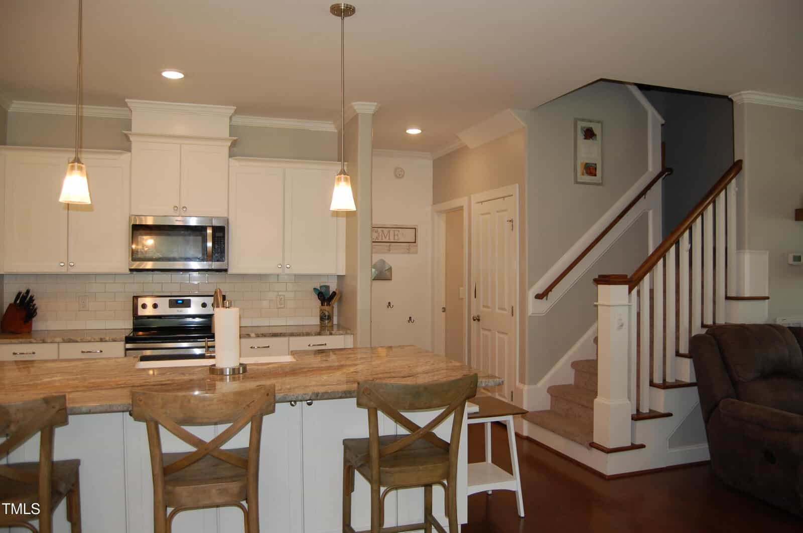 Modern kitchen with white cabinets, stainless steel appliances, granite island with three wooden chairs, pendant lights, and a nearby staircase with carpeted steps and white railing. Cozy, open-concept design.