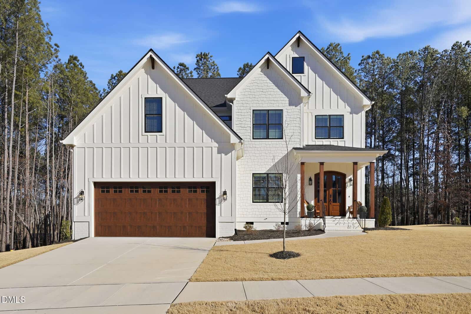 Two-story modern farmhouse with white siding, dark trim, a wooden double garage door, and a covered front porch with columns. Located at 100 N Ridge View Way in Franklinton, the house sits on a sloped lawn bordered by trees under a clear blue sky.