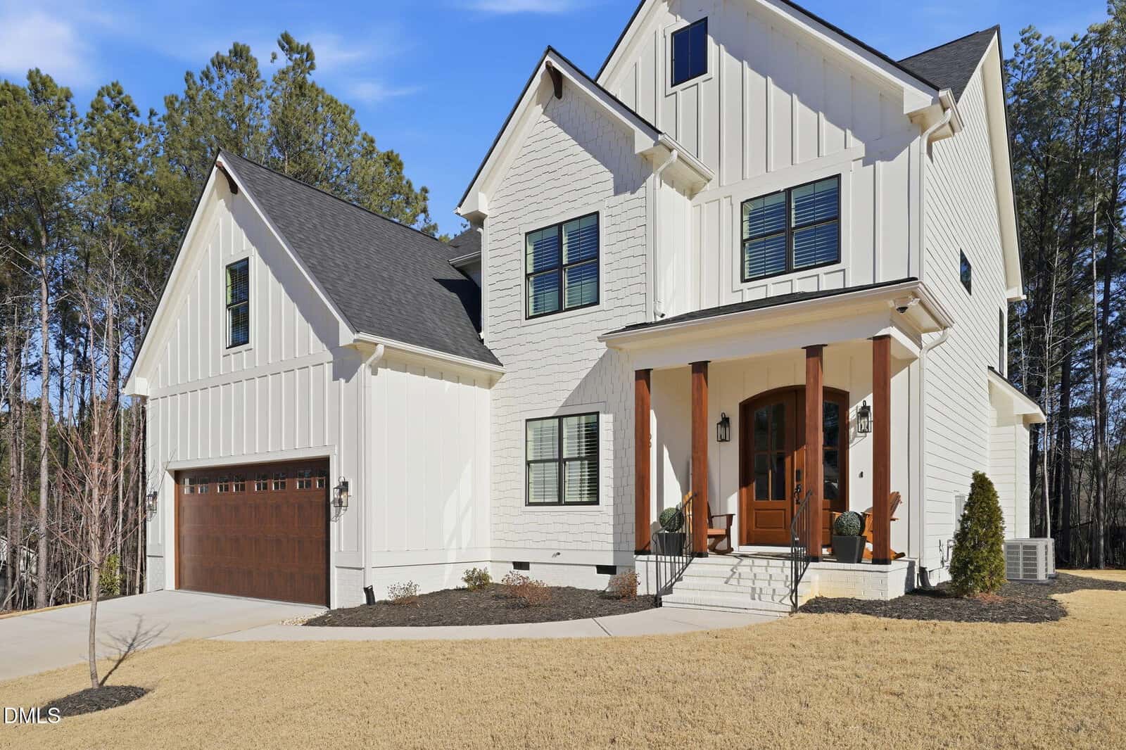 Two-story white house with black roof, wooden front porch columns, and a double garage with brown doors. The house sits on a lawn with minimal landscaping and trees in the background under a clear blue sky.