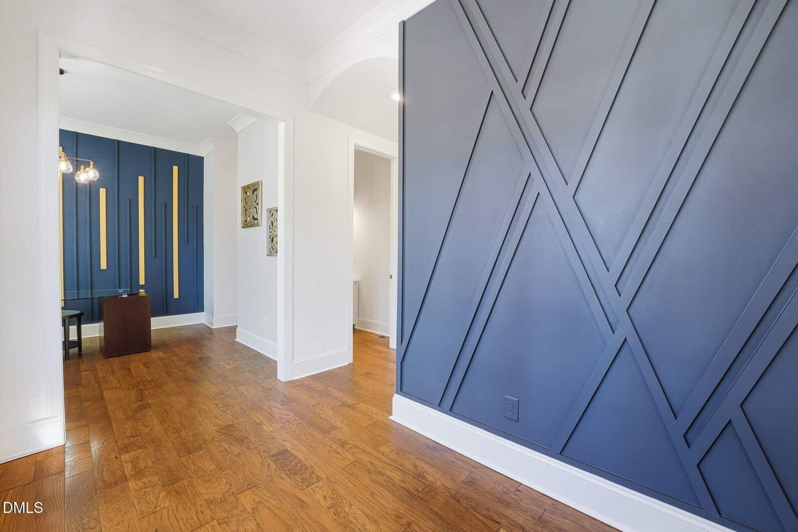 A modern interior with wood flooring and dark blue accent walls featuring geometric patterns. The space has white walls and trim, with a small table and chairs visible in an adjoining room. Natural light brightens the area.