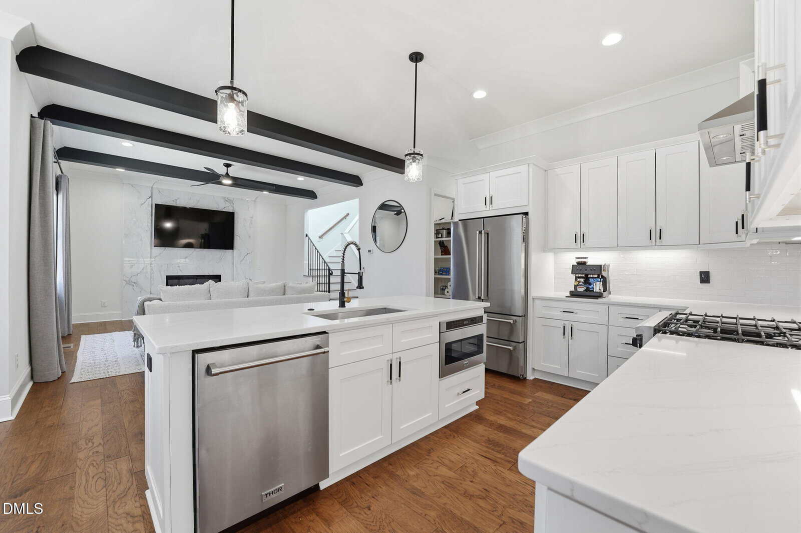 Modern kitchen with white cabinets, stainless steel appliances, an island with a sink, and hardwood floors. In the background, a living area with a TV above a fireplace and a circular mirror is visible. Black ceiling beams overhead.