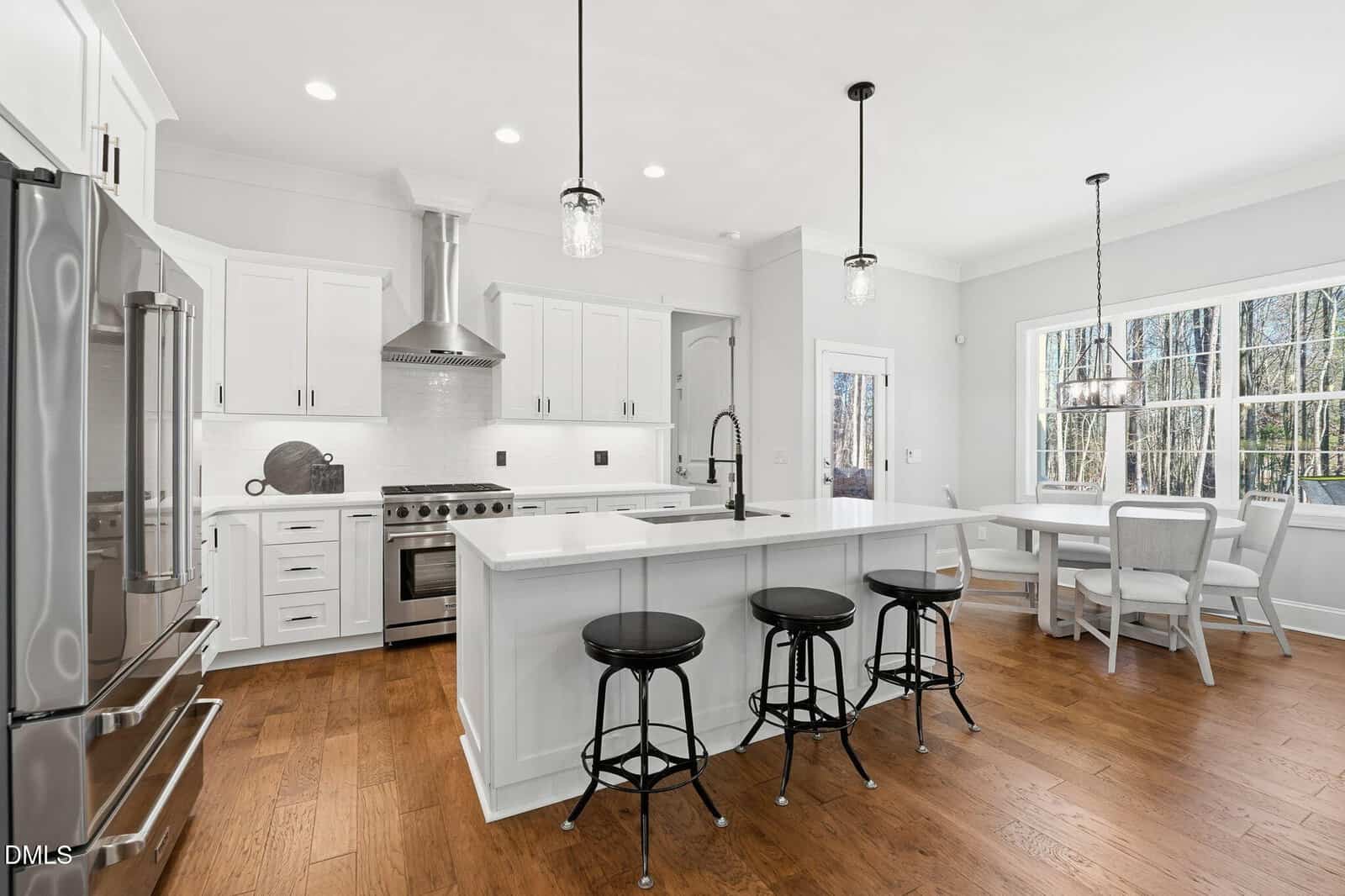 Modern kitchen with white cabinets, stainless steel appliances, a large island with three black stools, and a dining nook by large windows. Hardwood floors and pendant lights complete the bright, open space.