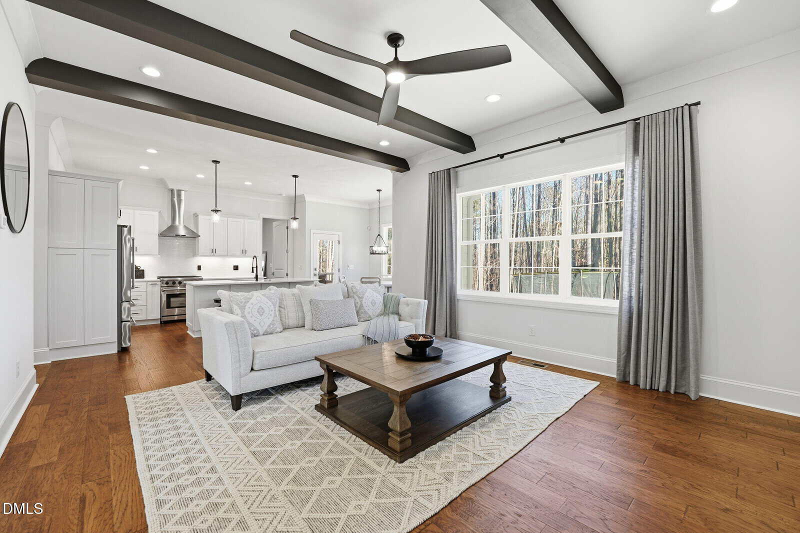 Modern living room with white walls, dark ceiling beams, a gray sofa, wooden coffee table, and large window with gray curtains. Kitchen with white cabinets and stainless steel appliances is visible in the background.