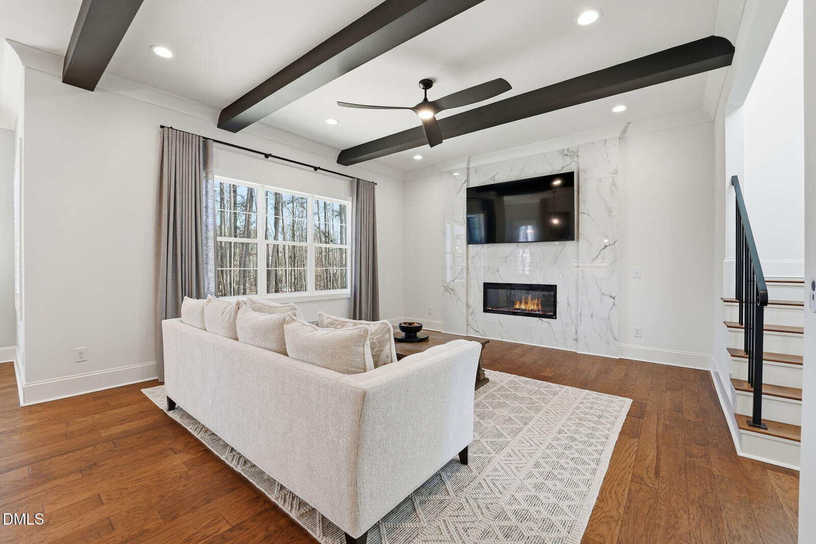 Modern living room with a white sofa, patterned rug, and wood floor. A wall-mounted TV and fireplace are framed by marble. Large window with gray curtains lets in natural light. Black ceiling beams and a ceiling fan are visible.