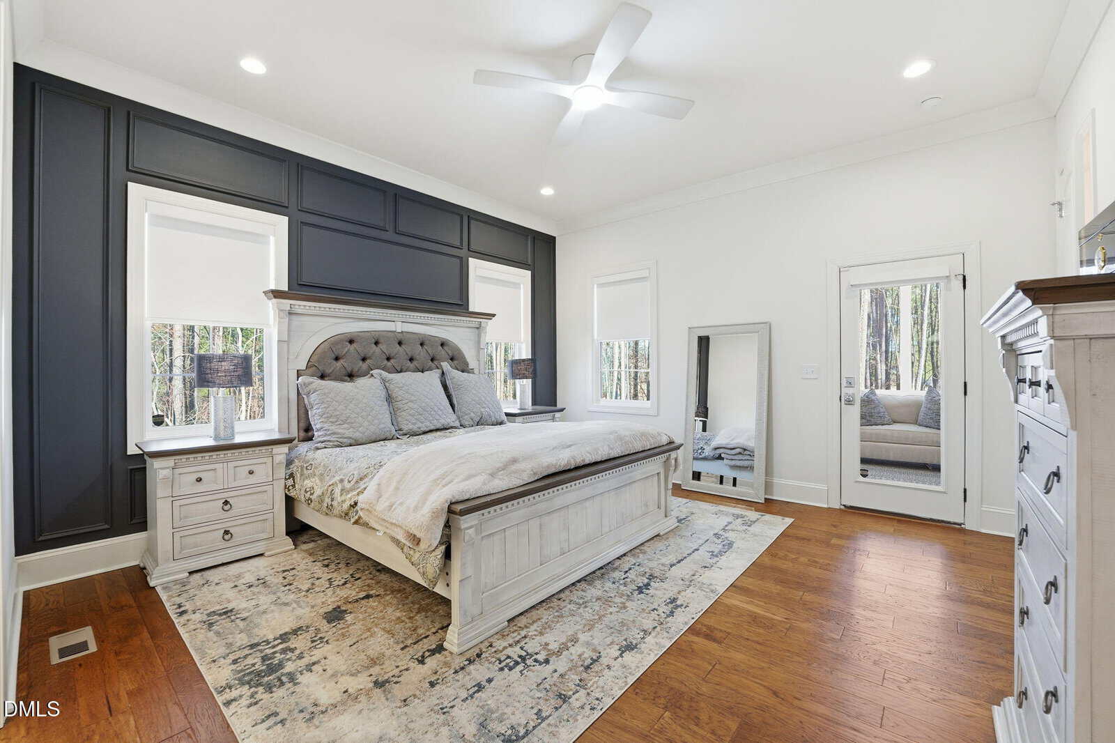 A bedroom with a large bed, two nightstands with lamps, a rug, wood flooring, a dresser, mirror, and windows with a view of trees. A sitting area is visible through a doorway in the reflection of the mirror.
