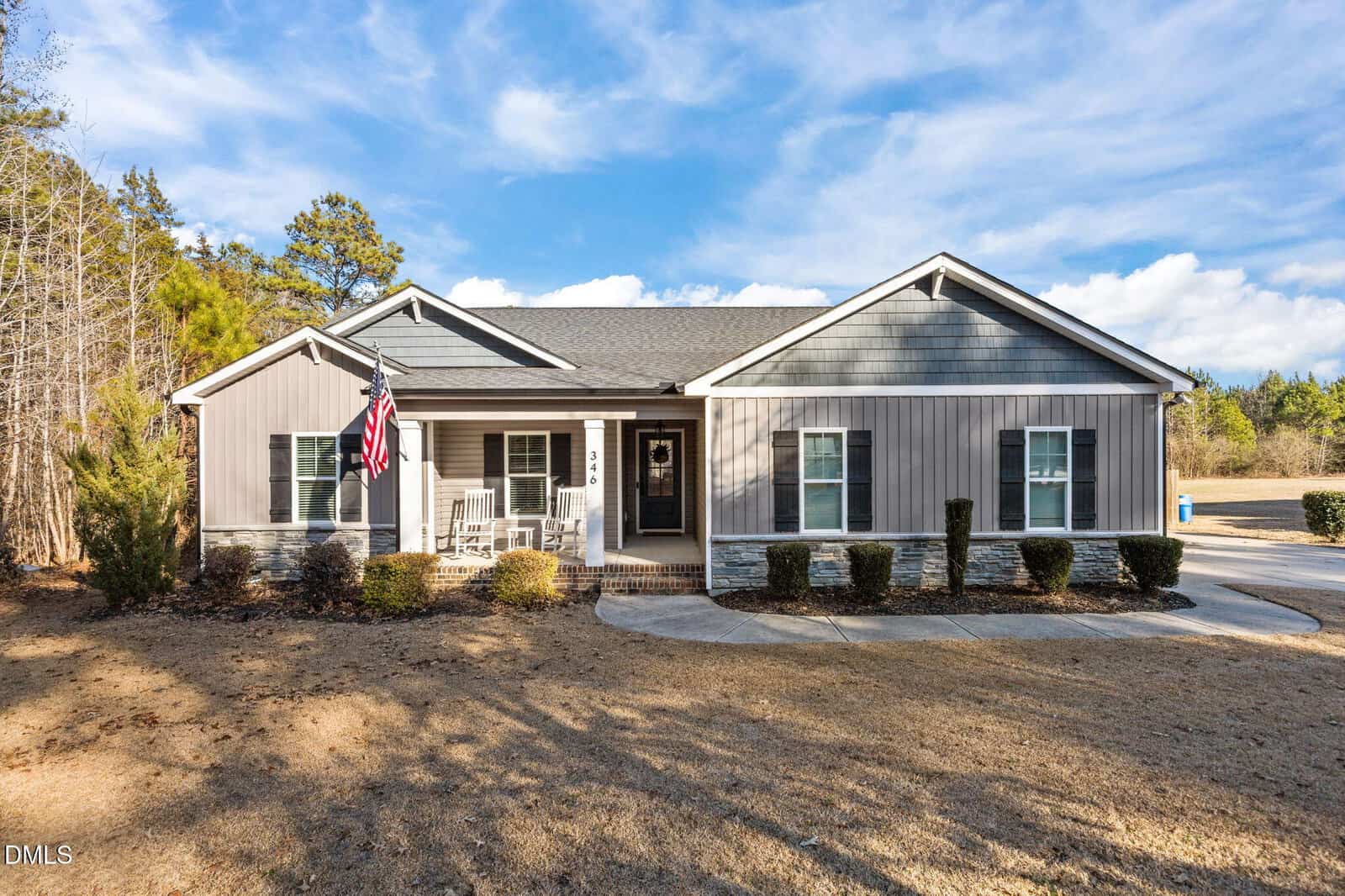Charming single-story gray house with white trim located at 346 Mount Olivet Church Road in Franklinton. Features a welcoming front porch, American flag, and neatly maintained shrubs in the front yard. The concrete driveway gently curves out front, while mature trees and a picturesque blue sky with clouds provide a scenic backdrop. The ground appears dry with some patches of grass.