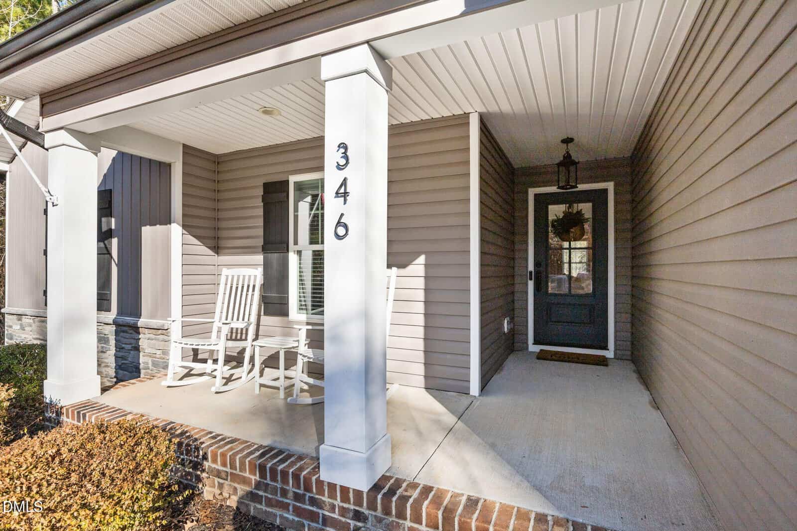 A front porch with two white rocking chairs, a black front door with a wreath, beige siding, a white column marked “346,” and a brick base. The porch floor is concrete and there is a lantern-style light above the door.