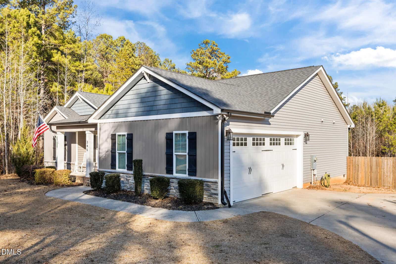 Single-story house with gray siding, white trim, and a two-car garage. There are shrubs along the front, an American flag by the porch, and a fenced backyard with trees in the background under a blue sky.