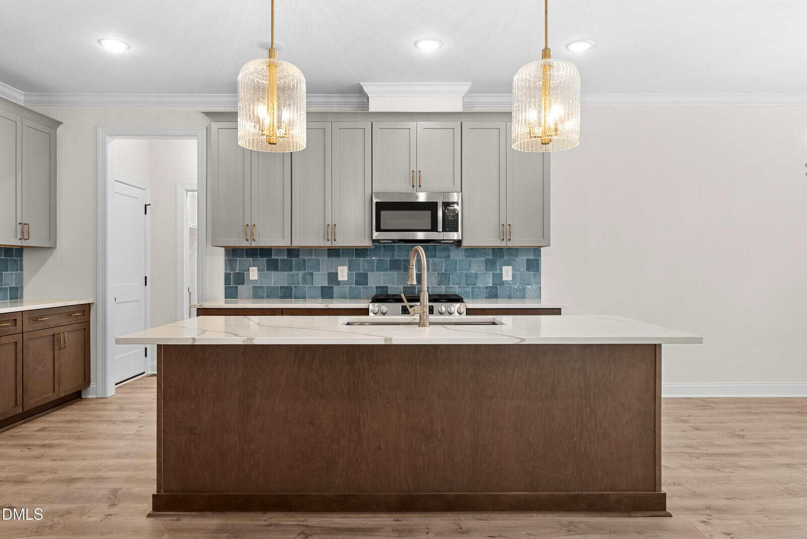 Modern kitchen with a large island, built-in sink, light brown cabinets, blue tile backsplash, stainless steel appliances, and two elegant pendant lights hanging over the island. Light wood flooring throughout.