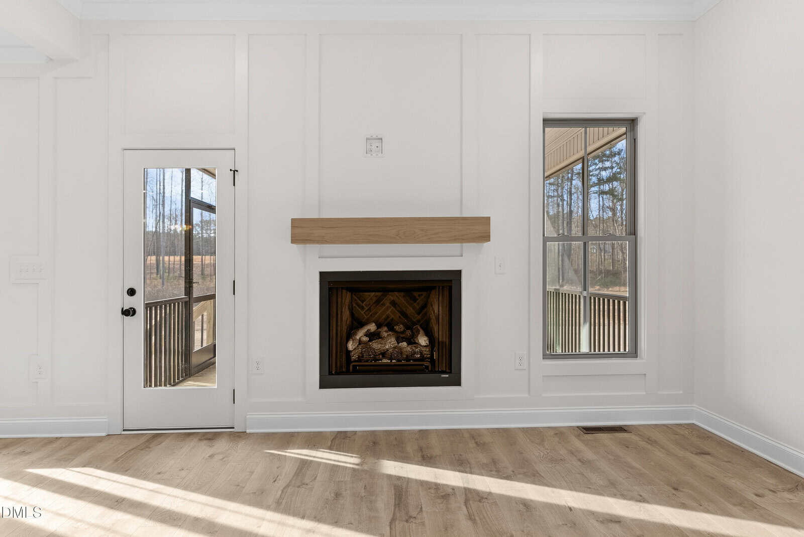 Bright living room with light wood floors, white walls, a modern fireplace with a wood mantel, large windows, and a glass door leading to an outdoor area. Trees and sunlight are visible outside.