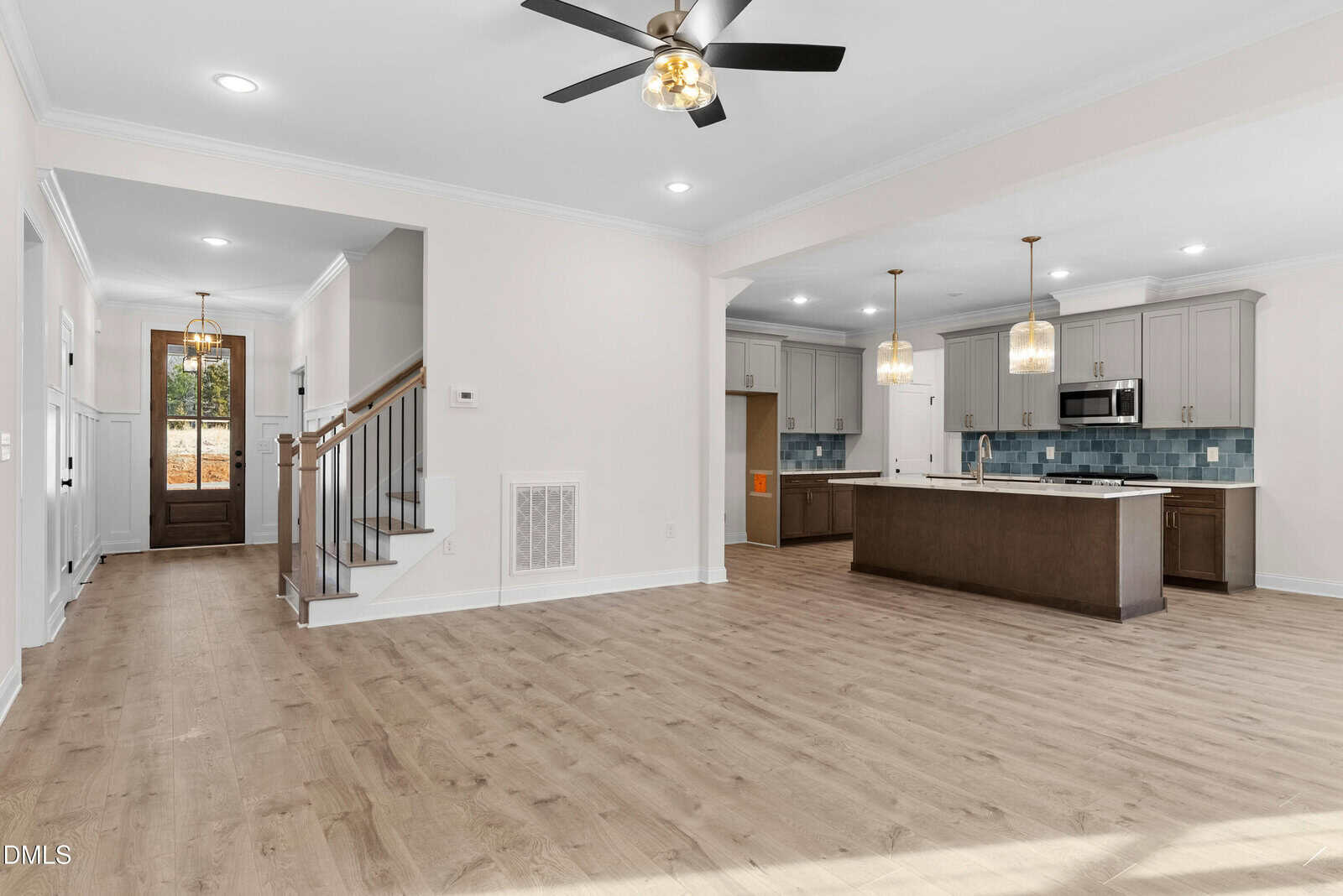 Modern open-concept kitchen and living area with light wood floors, gray cabinets, pendant lights over an island, staircase, and large front door in a bright, spacious home interior.