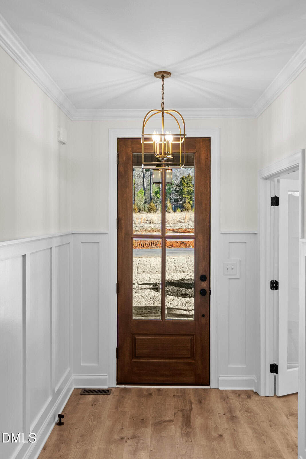 A hallway with white wainscoting, light wood floors, a wooden front door with glass panels, and a gold light fixture overhead. A window shows an outdoor view with trees and rocks.