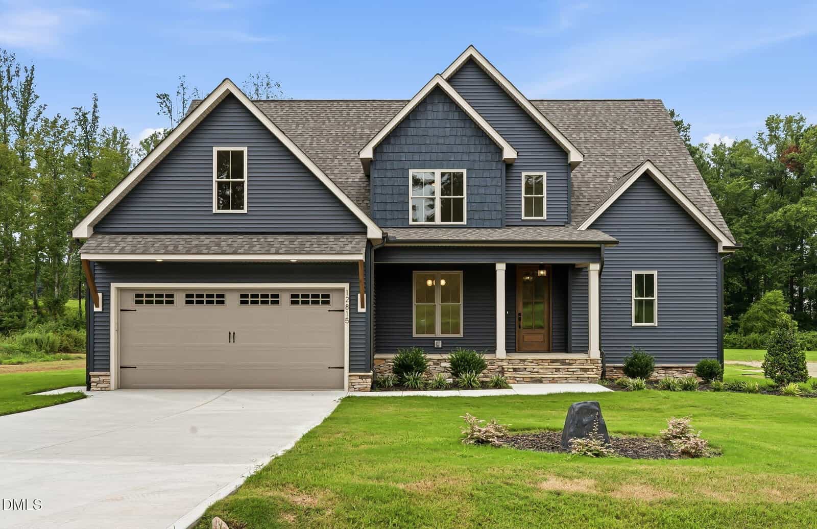 Modern two-story blue house with a gray shingle roof, two-car garage, covered porch, and landscaped front yard, set against a backdrop of trees and a blue sky. Situated at 12815 Cricket Cove Court in Zebulon, this residence offers both comfort and curb appeal in a picturesque setting.
