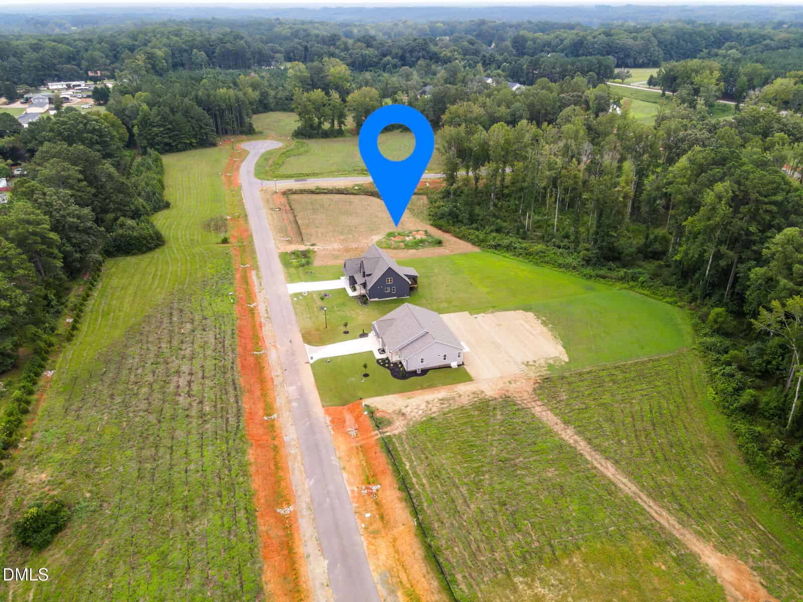 Aerial view of a rural neighborhood with two new houses, large green lawns, and a road. A blue location pin marks one house, surrounded by fields and trees.