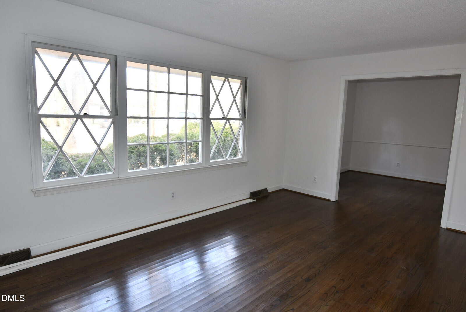 Empty room with large windows featuring diamond-patterned panes, white walls, and dark hardwood floors. An open doorway leads to another similar room. Some greenery is visible through the windows.