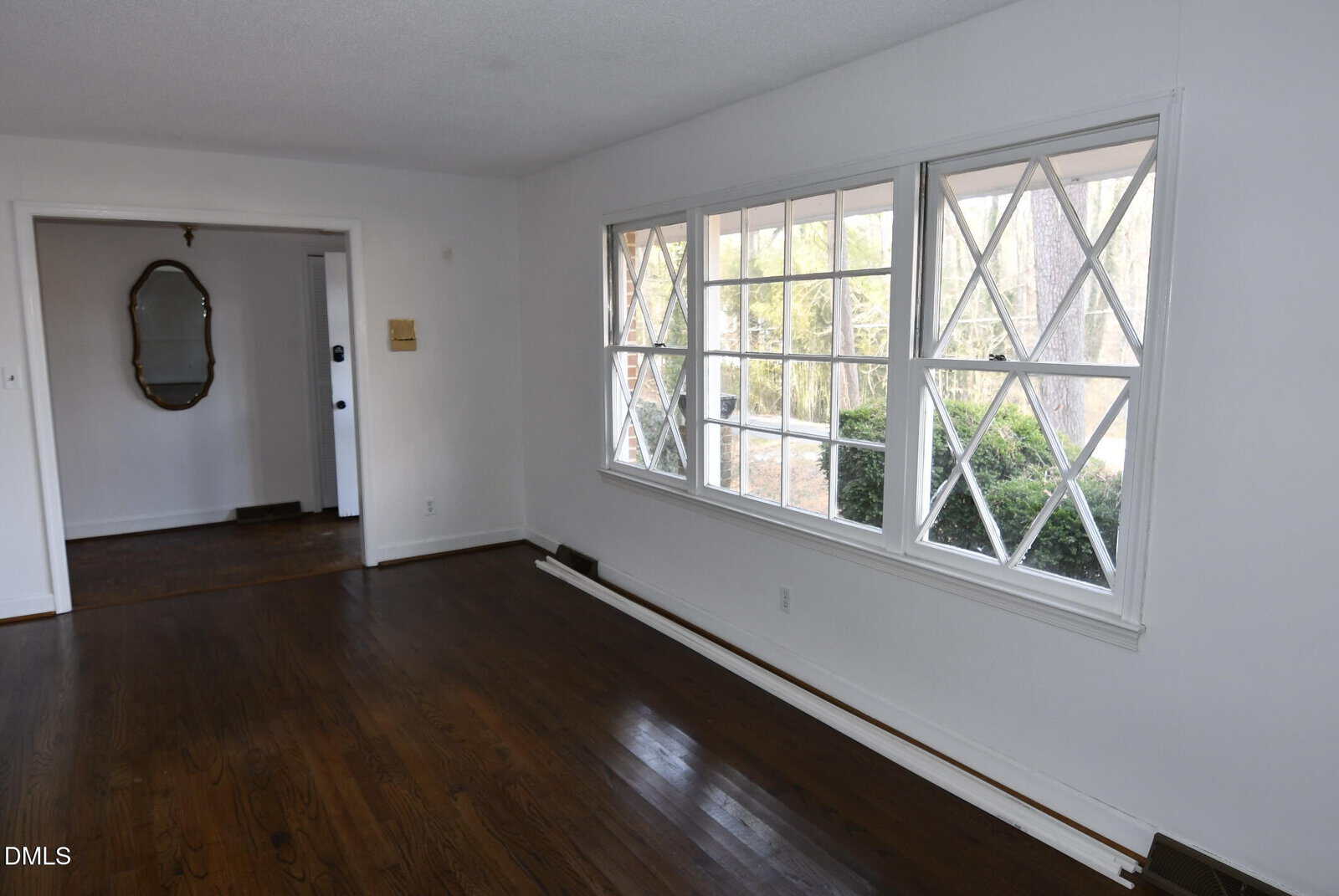 A room with dark wood floors, white walls, and a row of windows with diamond-shaped panes. An adjacent room with a wall mirror is visible through an open doorway. Shrubs are seen outside the window.