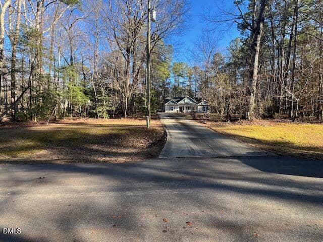 A long driveway leads through a wooded area to a white house with a front porch, set back from the road. Trees with sparse leaves surround the house under a clear blue sky.