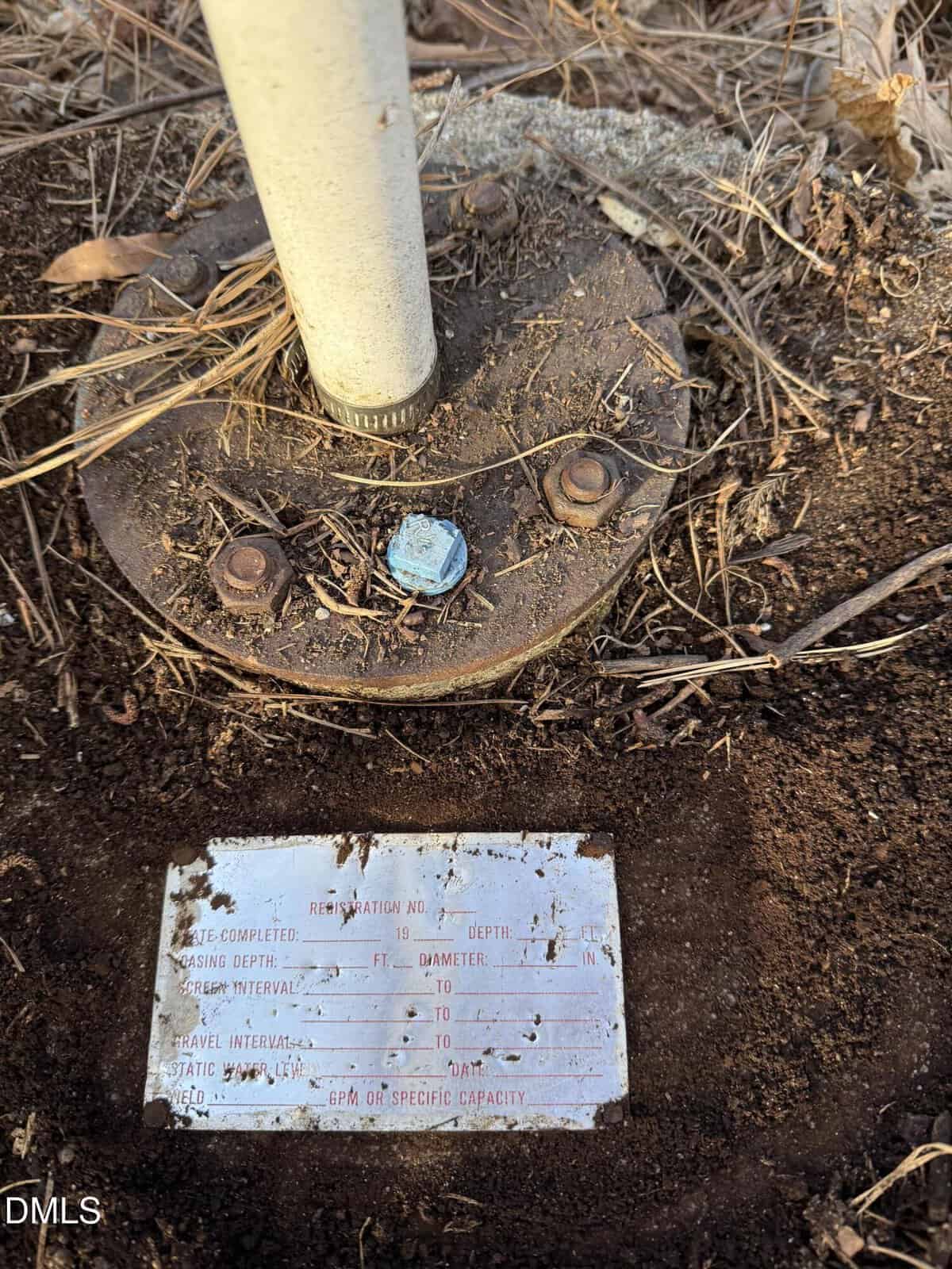 A metal pole is bolted to a round base surrounded by dirt and dry grass. Below the base, a weathered metal plate displays faded, hard-to-read engraved text with fields for depth, diameter, and intervals.