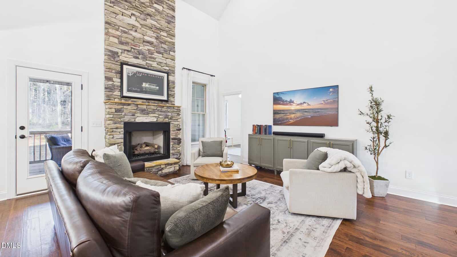 A living room with a stone fireplace, brown leather sofa, round wood coffee table, armchair, and TV on a cabinet. Hardwood floors, a rug, and a glass door leading outside are also visible.