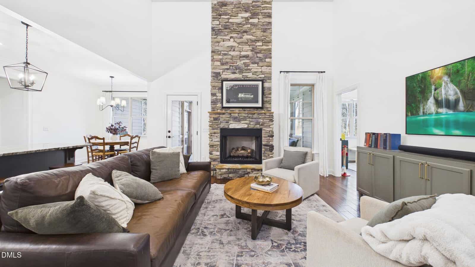 A living room with a stone fireplace, brown leather sofa, beige armchair, round wooden coffee table, TV on a gray cabinet, and a view of a dining area in the background. Decor is neutral and contemporary.