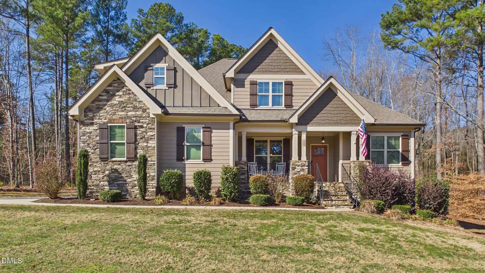 A two-story suburban house at 50 Willow Bend Drive in Youngsville, featuring a stone and siding exterior, multiple gables, a covered front porch with columns, landscaped bushes, an American flag, and surrounded by trees under a clear blue sky.