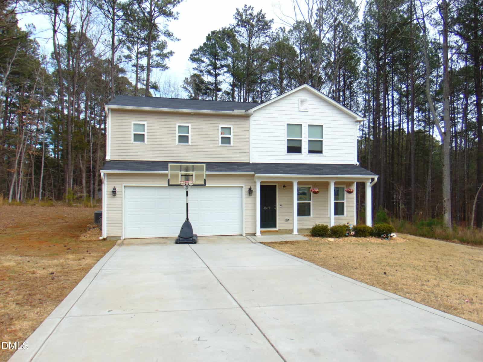 Two-story suburban house with a double garage, beige and white siding, a black front door, and a portable basketball hoop on the driveway. The yard has sparse grass and is bordered by trees.