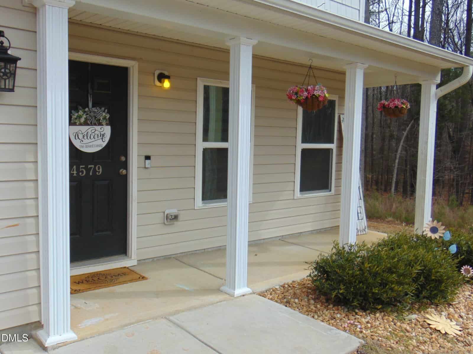A beige house with a black front door displaying a welcome sign, two white columns, two windows, a doormat, a lit porch light, hanging flower baskets, and shrubs near the entrance. House number 4579 is visible.