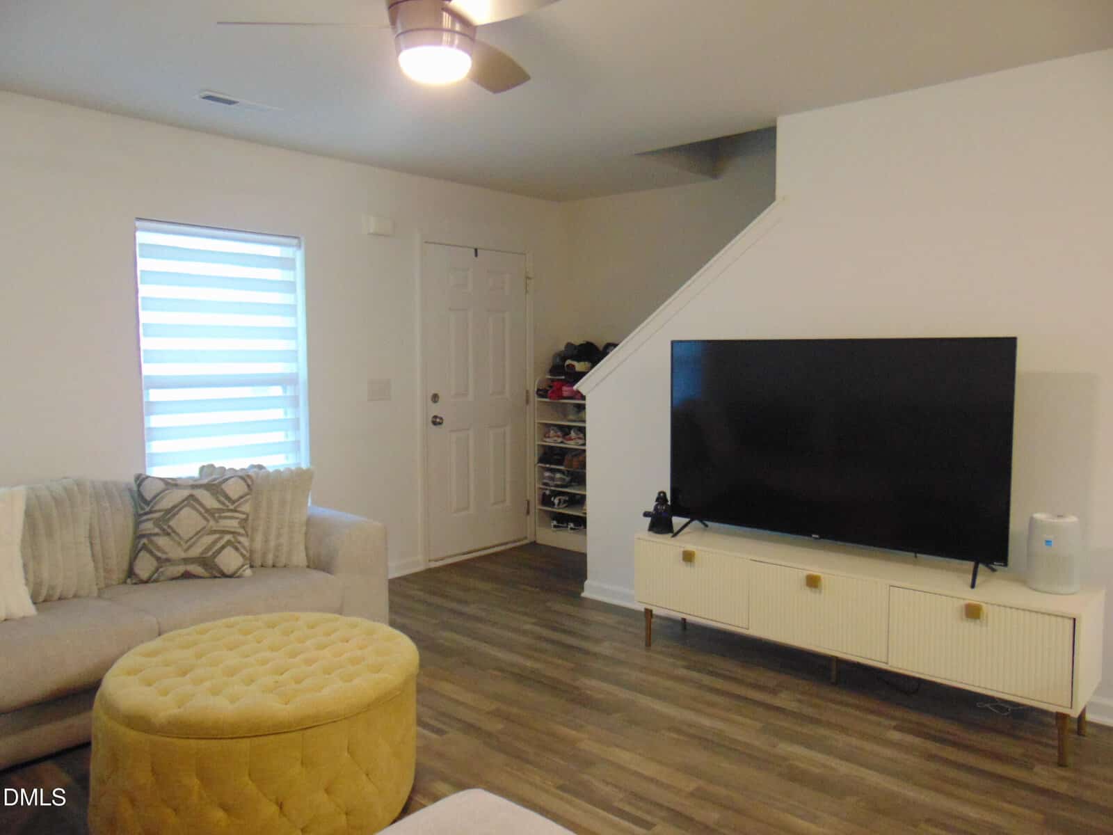 A living room with a gray sofa, patterned pillows, a round yellow ottoman, a large flat-screen TV on a white stand, wood flooring, a window with blinds, and a staircase near the front door.