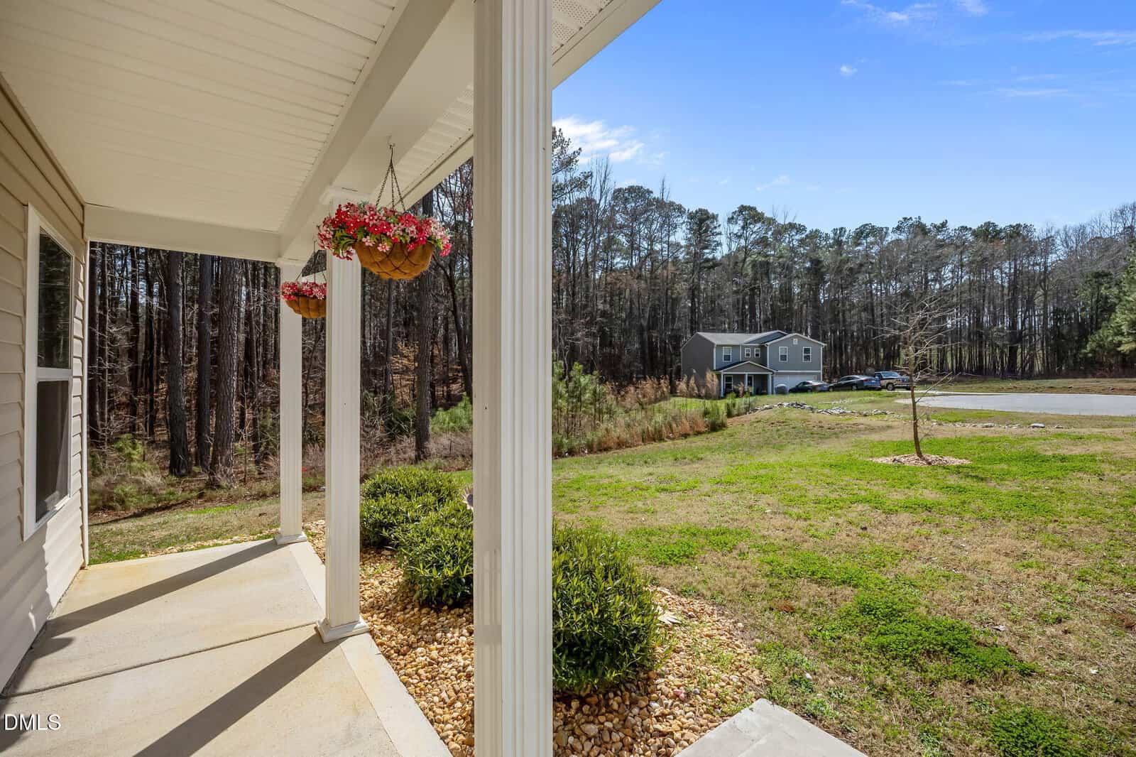 View from a porch with hanging flower baskets, looking out onto a grassy yard, driveway, and a two-story house across the road with trees in the background under a clear blue sky.